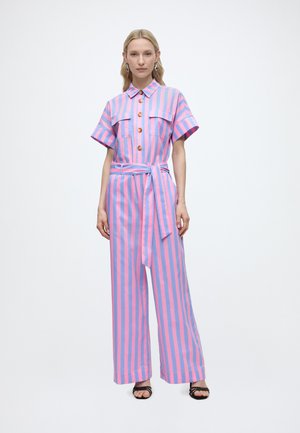 Femeie purtând un combinezon cu dungi verticale roz și albastre, cu mâneci scurte, nasturi în față, talie cu curea și sandale negre.