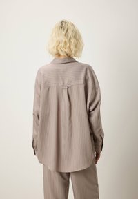 Persona con i capelli biondi corti indossa una camicia larga a righe verticali lilla chiaro e pantaloni abbinati, girata di spalle contro uno sfondo semplice.