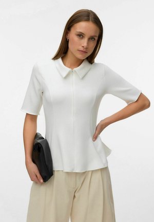 Jeune femme portant un haut blanc à manches courtes avec col et un pantalon beige, tenant une pochette noire, une main sur la hanche, devant un fond uni.