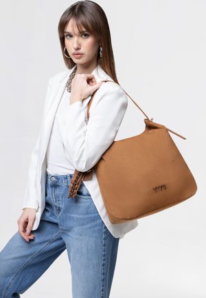 Joven mujer con blazer blanco y vaqueros sosteniendo un bolso de hombro de cuero marrón claro con logo sobre fondo claro.