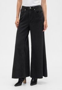 Jeans pretos de perna larga feitos de ganga, com cintura alta, dois bolsos frontais e acabamento desgastado. Combinados com saltos pretos pontiagudos.