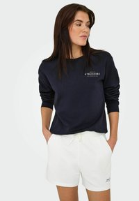 Mørkeblå sweatshirt med ribbede manchetter og hvidt logo; kombineret med hvide shorts med justerbar snøre og sidelommer.