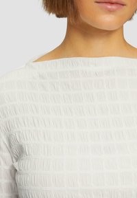 Blouse blanche texturée avec un motif froissé, dotée d'un décolleté large et d'un tissu lisse qui crée une apparence subtilement superposée.