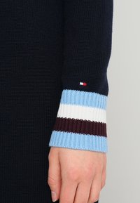 Manche de pull en maille marine avec un poignet côtelé rayé bleu, blanc et bordeaux. Petit logo près de la couture. Texture douce.