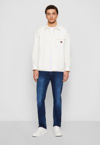 Νεαρός άνδρας στέκεται μπροστά σε γκρίζο φόντο, φορώντας ένα λευκό σακάκι, λευκό t-shirt, μπλε τζιν και λευκά αθλητικά παπούτσια.