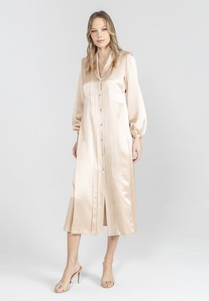 Kobieta stoi w beżowej satynowej sukience midi z długim rękawem i guzikami z przodu, w połączeniu z beżowymi, odkrytymi na palcach klapkami na wysokim obcasie.