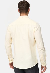 Lys gul langærmet skjorte med knap-krave, tekstureret stof og knaplukninger ved manchetterne. Bagvæggen viser lige kant og sømme.