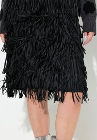 Jupe à franges noires en tissu texturé, présentant de longues bandes pendantes et un ourlet juste au-dessus du genou. Chaussures argentées visibles.