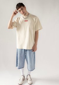 Beige oversized t-shirt with red "WARNING" text, paired with blue denim shorts, white socks, and brown/white sneakers. Silver chain accessory.