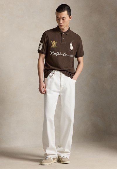 Νέος άνδρας με καφέ polo Ralph Lauren με λογότυπα, λευκά τζιν με φαρδιά πόδια και μπεζ αθλητικά παπούτσια, στέκεται με το ένα χέρι στην τσέπη.