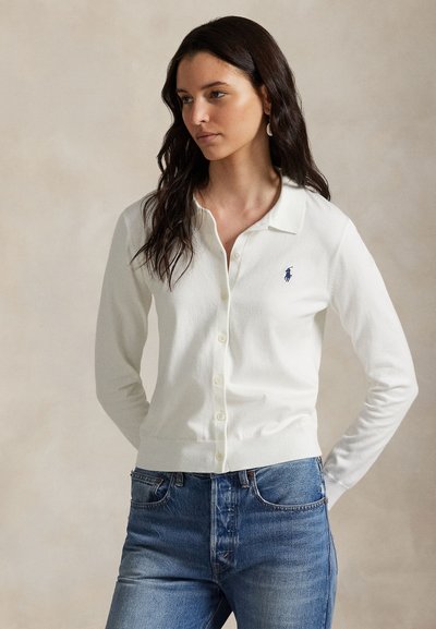 Bel beli kardigan iz mehkega blaga z osnovnim ovratnikom, dolgimi rokavi in majhno modro majhno modro značko na levi strani prsnega koša.