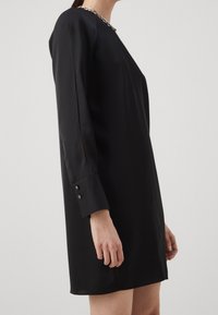 Frau in einem schwarzen Langarmkleid mit geknöpften Manschetten und einer silbernen Kettenhalskette, von der Seite vor einem schlichten Hintergrund gezeigt.