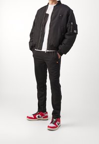 Zwarte bomberjack met geribbelde manchetten en kraag, wit T-shirt, zwarte broek met bies, en rood-grijze sneakers met zwarte accenten.