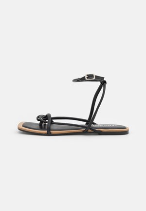 Sandalia plana negra con tiras finas, diseño con nudo, correa ajustable en el tobillo y suela minimalista con detalles en colores claros.