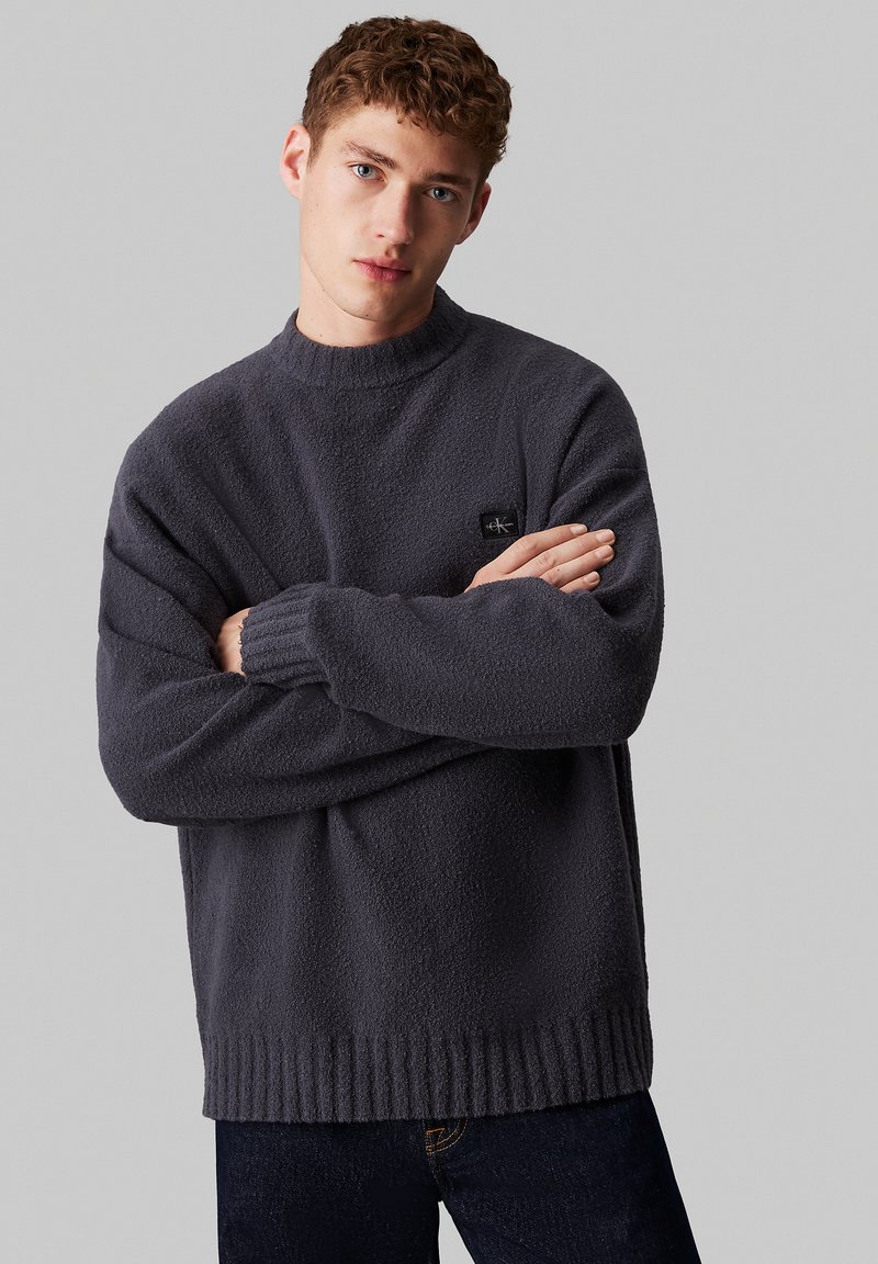 Maglione in pile grigio scuro con collo alto, orli a costine e maniche morbide. Presenta una piccola etichetta con logo sul petto.