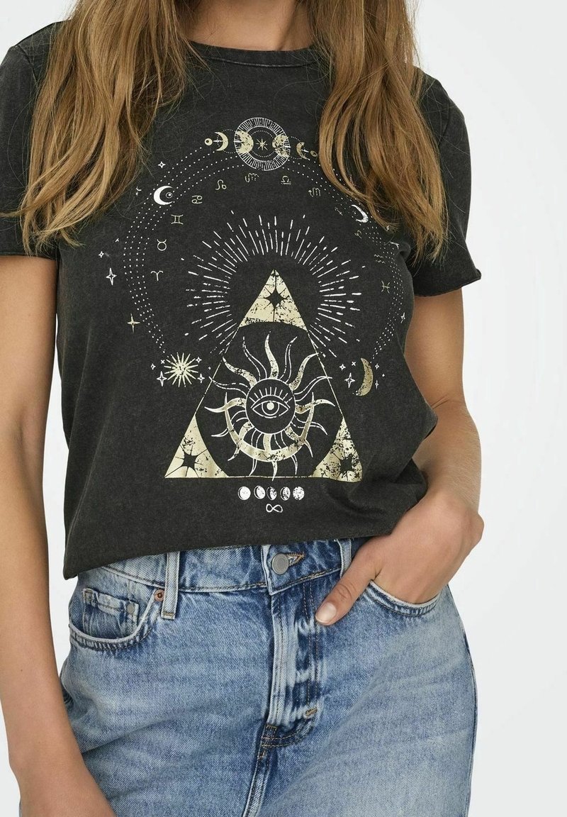 Femme portant un t-shirt noir avec un motif cosmique incluant un œil dans un soleil à l’intérieur d’un triangle, des phases de la lune et des symboles du zodiaque.