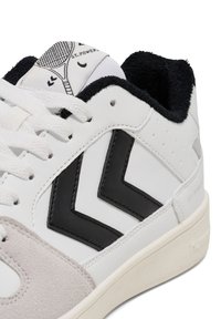 Witte sneaker met zwart chevrondesign, grijze suède accenten, gestructureerde zwarte kraag en een logo op de tong met een tennismotief.