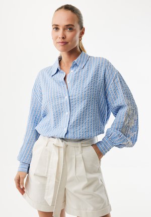 Lichtblauw overhemd met verticale witte strepen, knoopsluiting, losse pasvorm en geborduurd bloemmotief op de mouwen. Gecombineerd met witte shorts.