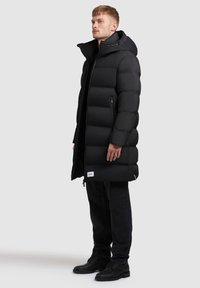 Long black puffer coat with a hood, featuring quilted sections, zippered side pockets, and a logo patch on the lower front. Textured fabric.