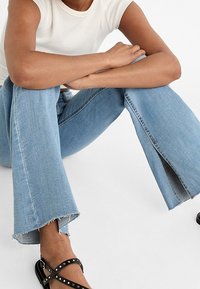 Lichtblauwe denim flared jeans met raw hems en zijsplits, gecombineerd met zwarte sandals met bandjes en metalen accenten.