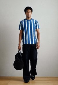 Camiseta de manga curta às riscas azul e branca, calças de ganga pretas largas, segurando uma mala redonda preta, em pé num chão de madeira contra um fundo neutro.