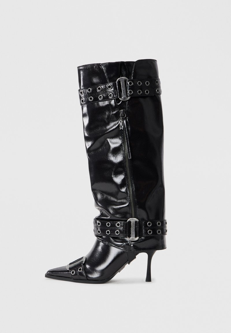 Botte en cuir verni noir montée jusqu'au genou, avec un bout pointu, une sangle à la cheville dotée de boucles métalliques et une fermeture éclair sur le côté. Texture lisse avec une finition brillante.