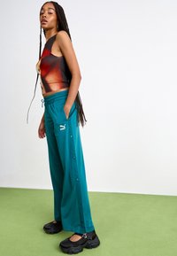 Teal viddebeinte bukser med sideknapper, sammen med en svart ermeløs topp med et livlig grafisk design. Svart chunky sko.