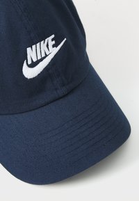 Tamna plava kapa od tkanine s zakrivljenim pokrivačem, s bijelim izvezenim Nike logotipom na prednjoj strani. Tekstuirani materijal, jedinstvena boja.