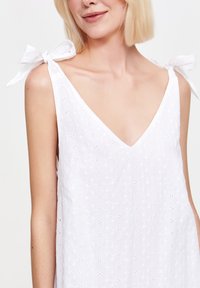 Zbliżenie na kobietę z krótkimi blond włosami, ubraną w białą sukienkę bez rękawów, ozdobioną materiałem w dekoracyjne wzory i kokardkami na ramionach.