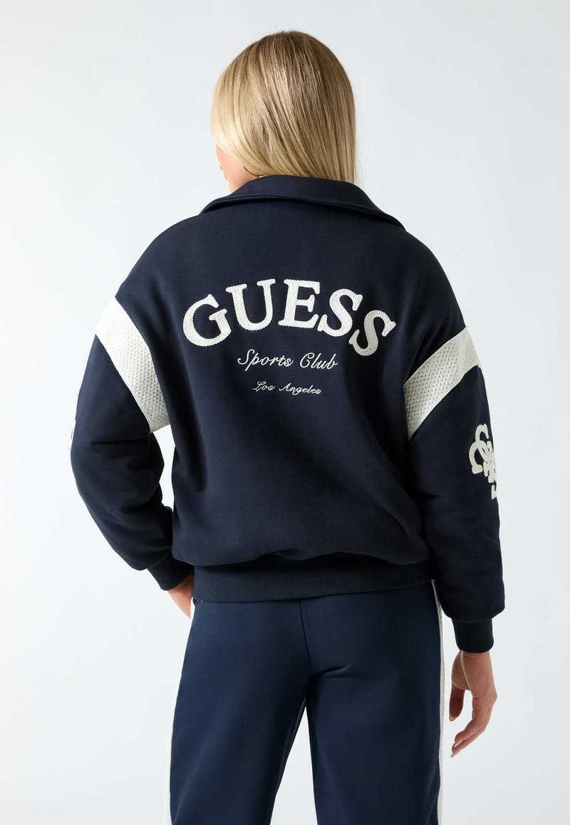 Personne blonde portant une veste de sport bleu marine avec un texte blanc "GUESS Sports Club Los Angeles" dans le dos et un pantalon assorti.