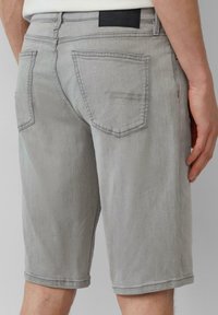 Des shorts en denim gris présentent une coupe droite, avec des poches au dos et une étiquette de taille noire. Texture lisse avec de subtils détails de couture.