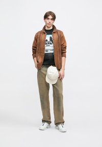 Καφέ πουλόβερ με υφή, μαύρη μπλούζα με γραφικά, ανοιχτό πράσινο παντελόνι και λευκά αθλητικά παπούτσια. Ένα κρεμ καπέλο κρατιέται στο αριστερό χέρι.