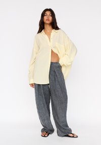 Světlé žluté oversized košile s širokými rukávy, spárované s temně šedými lněnými kalhotami s širokými nohavicemi. Celý outfit doplňují ploché černé sandály.