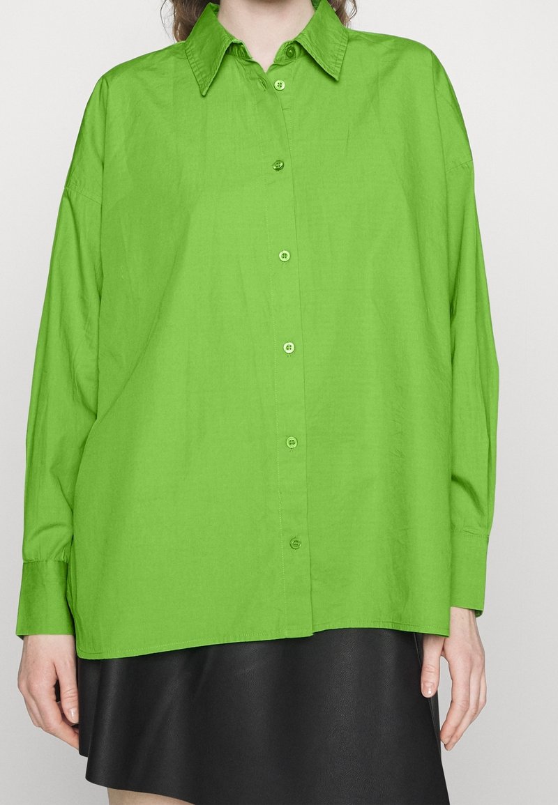 Femme portant une chemise oversize vert vif à manches longues et une jupe noire, debout devant un fond uni.