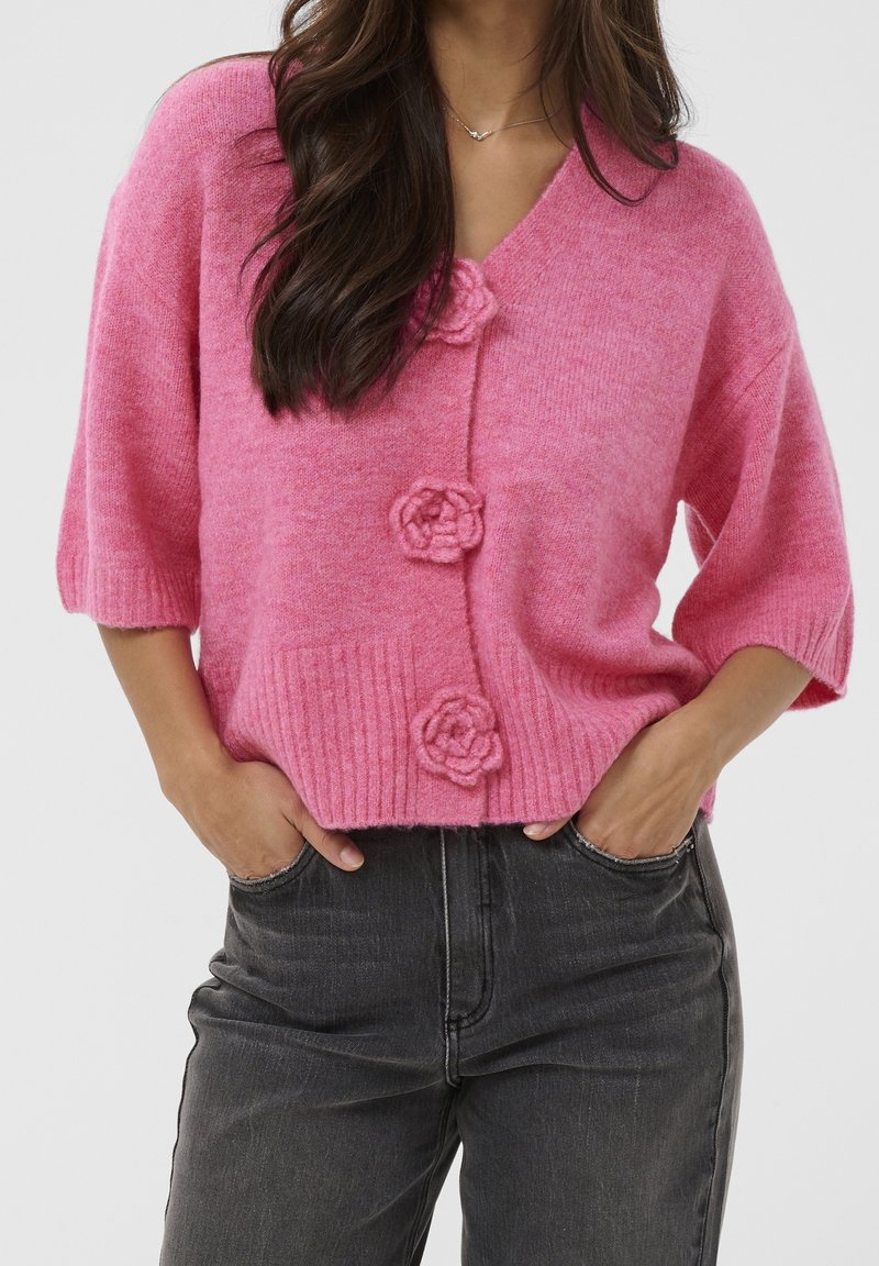 Femme portant un cardigan rose duveteux avec trois boutons en forme de fleur et un jean noir taille haute, mains dans les poches, sur un fond uni.
