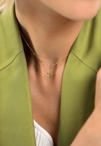 Złoty naszyjnik z krzyżykiem na cienkim łańcuszku. Noszony na zielonym blezerze, teksturowana tkanina kontrastuje z gładkim metalem.