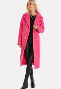 Cappotto lungo doppiopetto in tessuto rosa brillante, con grandi bottoni e ampi revers. Indossato sopra un top nero e pantaloni in pelle.