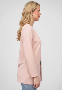 Lichtroze lange mouwen top met horizontale strepen, ronde halslijn en een relaxed fit, gecombineerd met blauwe denim jeans. Zijaanzicht.