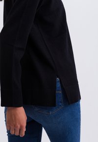 Schwarzer Strickpullover mit seitlichen Schlitzen und gerippten Bündchen, kombiniert mit blauen Jeans. Weicher Stoff und taillierte Passform, die lässige Mode betont.