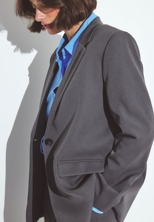 Zwarte op maat gemaakte blazer met een enkele knop, voorzien van een gladde stof, scherpe revers en zijzakken, gelaagd over een blauwe blouse.
