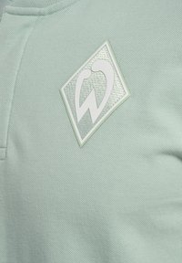 Miętowa, teksturowana tkanina z prostokątną łatką z marką, na której znajduje się biały projekt logo, umieszczoną w górnej części ubrania.