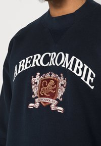 Námořnická mikina s bílou výšivkou "ABERCROMBIE", s červeným a zlatým erbem s tygrem a textem "NEW YORK, NY".
