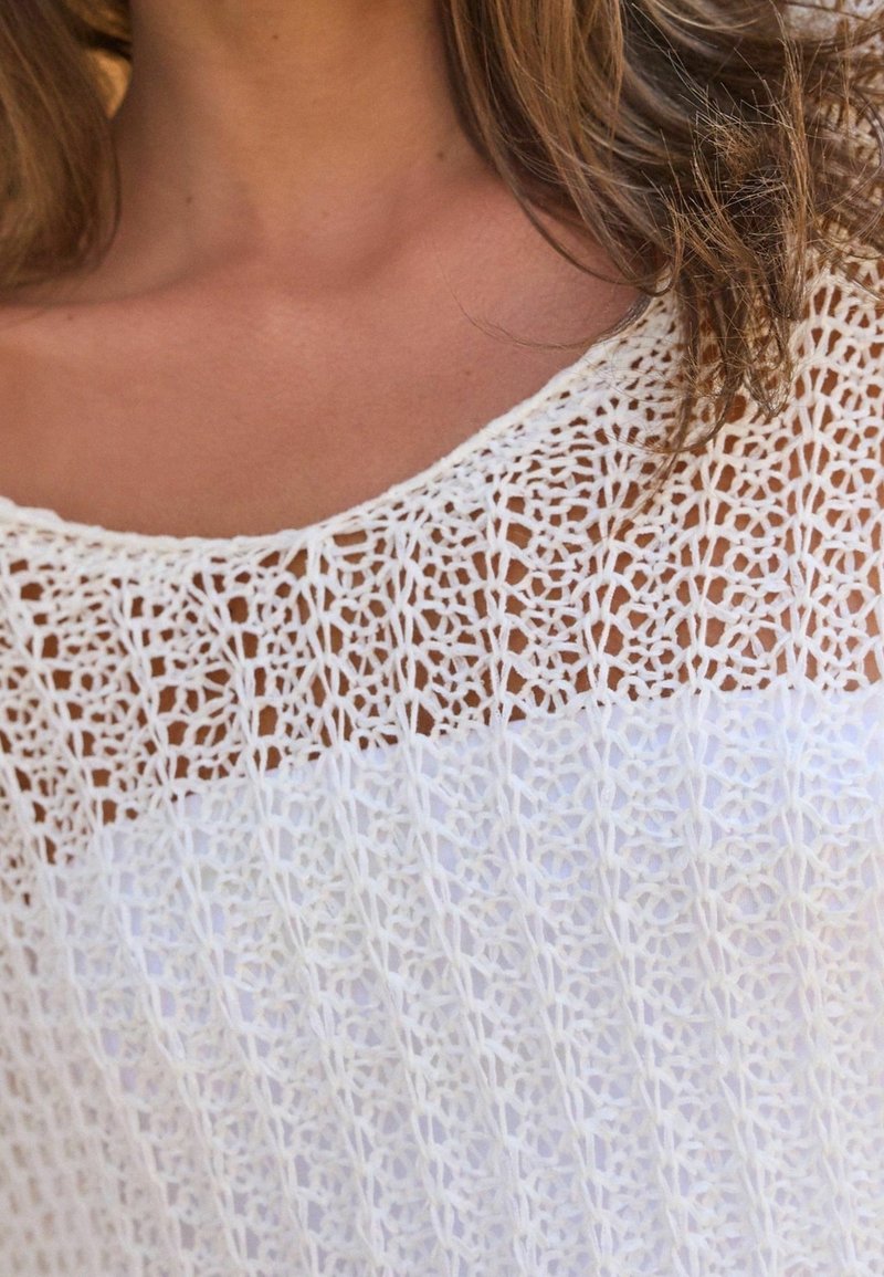 Haut tricoté blanc avec un motif ajouré et une section blanche unie en dessous. Encolure ronde et tissu léger texturé.