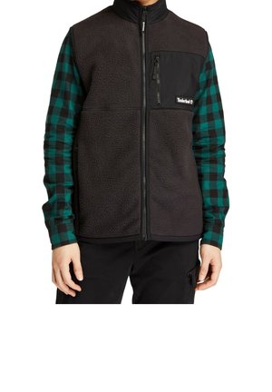 Personne portant un gilet en polaire noir avec une poche poitrine zippée sur une chemise à carreaux verts et noirs à manches longues et un pantalon noir.
