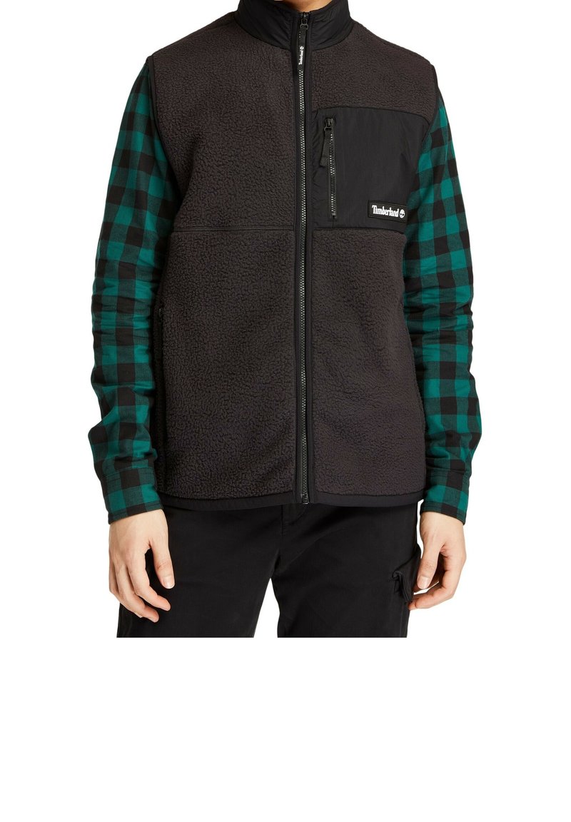 Personne portant un gilet en polaire noir avec une poche poitrine zippée sur une chemise à carreaux verts et noirs à manches longues et un pantalon noir.