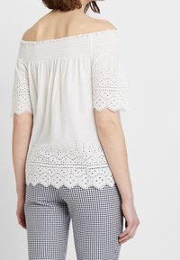 Top blanco sin mangas con escote smock, patrones de ojales en las mangas y el dobladillo, combinado con pantalones de cuadros en blanco y negro.