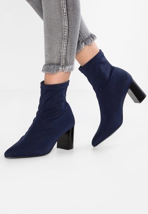 Person iført marineblå sokkestøvler med spids tå og blokhæl og grå jeans med frynset kant mod en hvid baggrund.