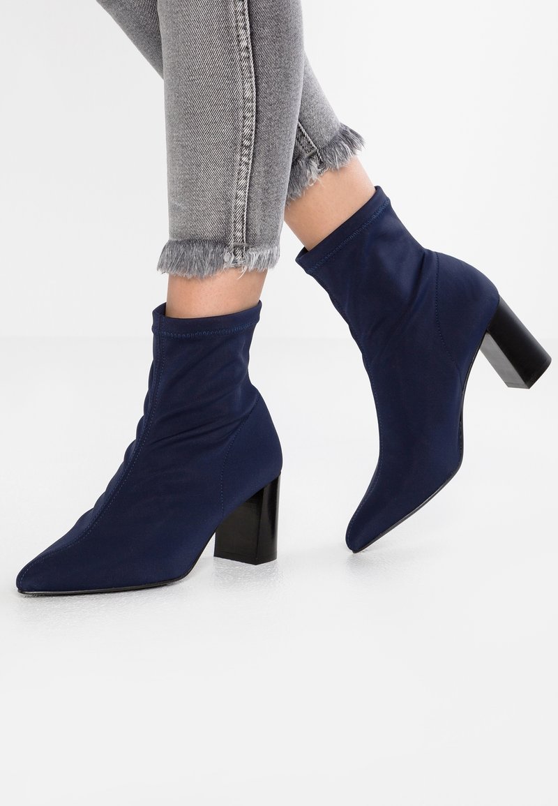Personne portant des bottines chaussettes bleu marine à bout pointu avec talons blocs et un jean gris à ourlet effiloché sur fond blanc.