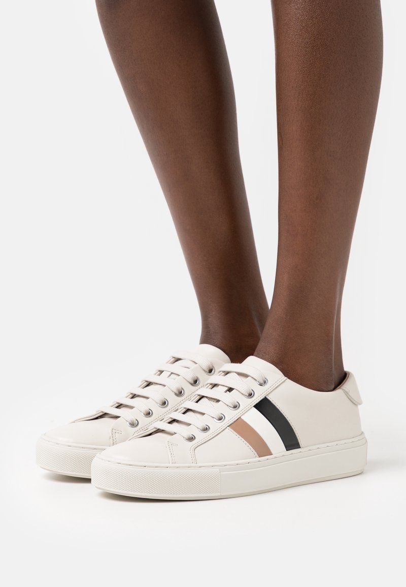 Baskets en cuir beige avec des bandes noires et marron, bout rond, semelle plate et œillets en métal. Les lacets se nouent à l'avant.