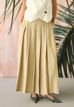 Frau trägt beige plissierte Hosen mit weitem Bein, weiße ärmellose Weste und Zebra-musterte Kitten Heels, steht vor einem tropischen Blätterhintergrund.